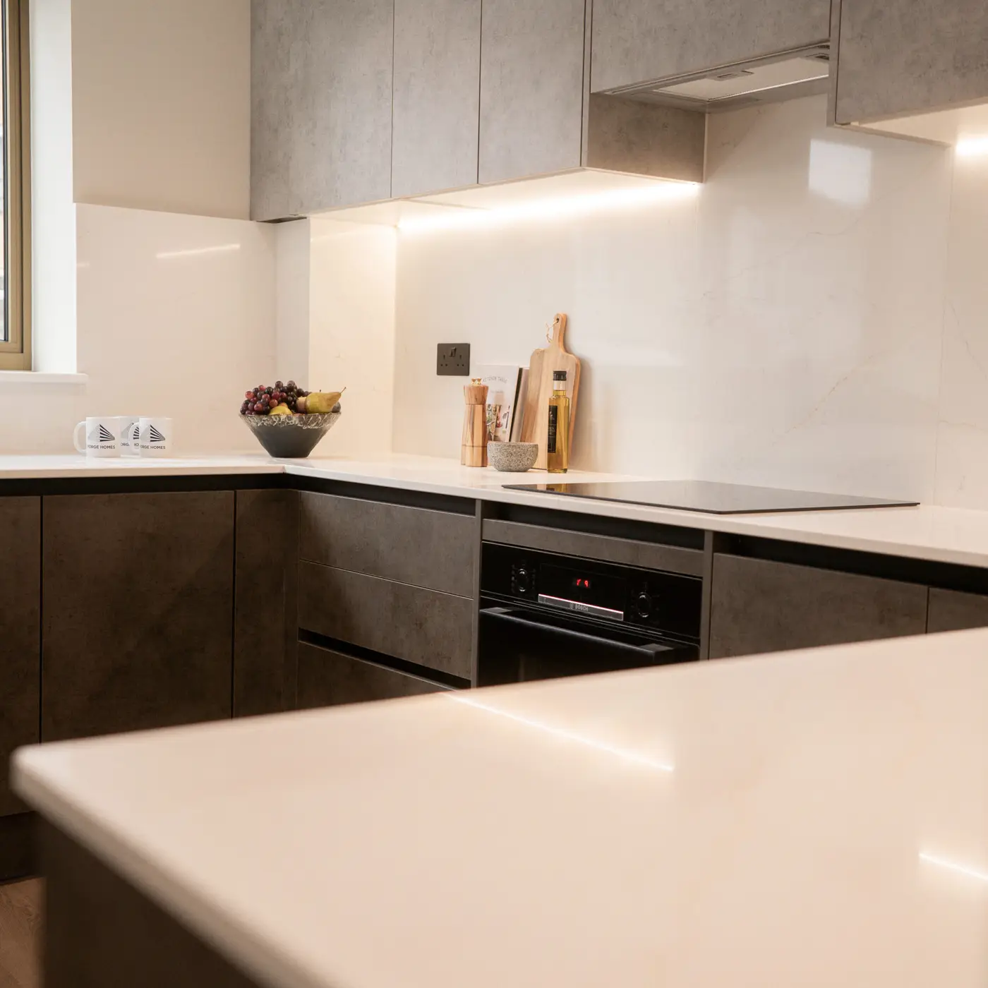 Light slate grey top cabinets with white marble splashback and countertops. Led lights underneath the grey top cabinets. Decorative items on top of worktop, with mugs, fruit and countertop, oils, and other cooking utensils