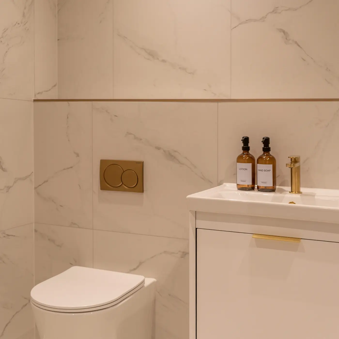 Shot of the main bathroom with the floating white sink basin with gold features. White marble tiling and gold features. White toilet